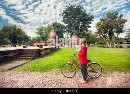Woman in red shirt with bicycle looking at old ruined Buddhist temple in Sukhothai historical park, Thailand Stock Photo