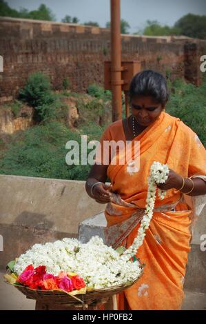 Indian woman in orange sari threading jasmine flowers into a garland outside a temple, Tamil Nadu, South India Stock Photo