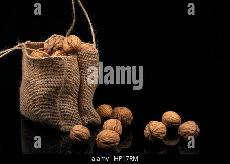 Walnuts in the bag of burlap. Spilling nuts lie on a glossy surface on a black background Stock Photo