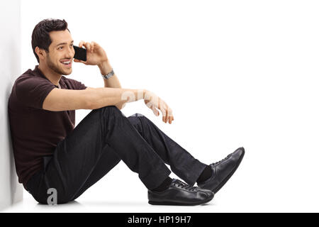 Young man talking on a phone while sitting on the floor and leaning against a wall isolated on white background Stock Photo