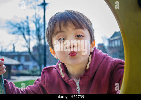 A young boy poses for a photo through a porthole in a climbing frame in a children's playground. Stock Photo