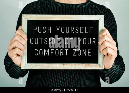 closeup of a young caucasian man holding a chalkboard in front of him with the text push yourself outside of your comfort zone written in it Stock Photo
