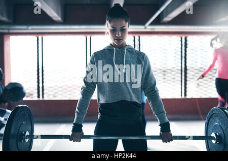 Shot of young pretty woman in sportswear training in gym and lifting barbell against barred window. Stock Photo