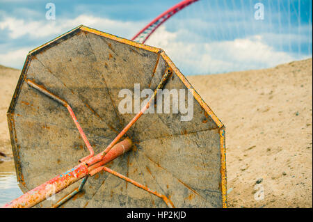 abandoned rusty metal parasol on the sandy beach, on a background of red bridge and blue sky Stock Photo