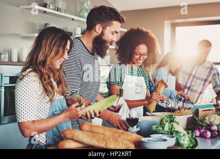 Good friends laughing and talking while preparing meals at table full of vegetables and pasta ready for cooking in kitchen Stock Photo
