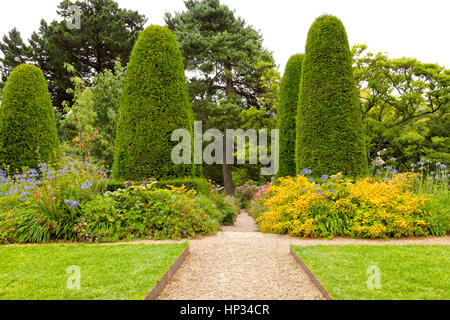 Stone pathway crossing summer flowers flowerbeds and lawn in a landscaped garden, with pine trees and shaped conifers Stock Photo
