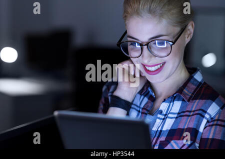 Amused IT girl using gadget in the dark lighted room Stock Photo