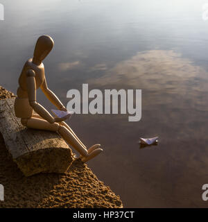 Wooden man is sitting on the beach and launching paper boats Stock Photo