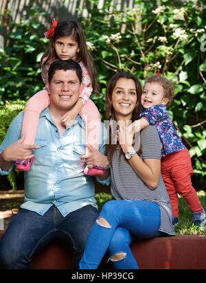 Young latino family of four in green park background sitting on brench Stock Photo