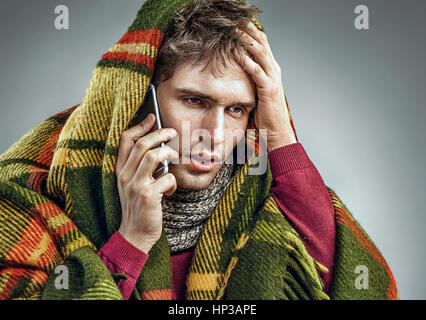 Unhealthy man wrapped in blanket with high temperature calling on the phone. Man suffering cold and winter flu virus Stock Photo