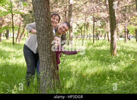Happy family and child in summer park. People hiding and playing behind a tree. Beautiful landscape with trees and green grass Stock Photo