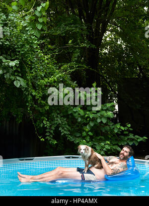 A miniature Australian Shepard sitting on a man and his floatie in a pool. Stock Photo