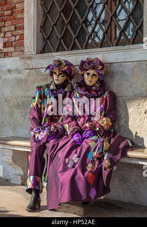 Venice, Italy- February 18th, 2012: Two people disguised in purple Venetian costumes sitting on a bench during the Venice Carnival days. Stock Photo