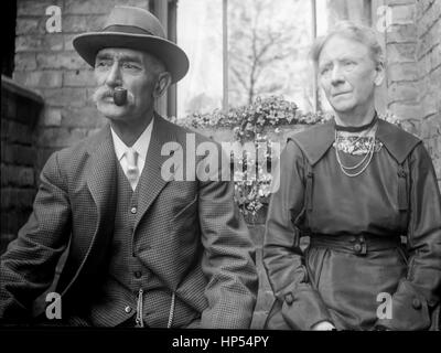 Elderly Victorian couple sitting outside a house with a flower box and curtained window in the background. The man has a mustache and  dressed in a suit and tie with a watch chain across his waistcoat. The lady is wearing a long, plain, black dress.  Photograph taken 1900, restored from a high resolution scan taken from the original negative. Stock Photo