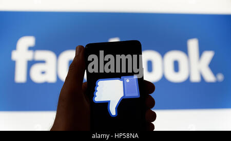 Hand holding a mobile phone with a dislike button picture on it. Facebook's logo is shown on the background in a monitor. Stock Photo