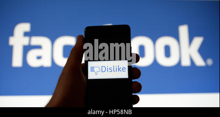 Hand holding a mobile phone with a dislike button picture on it. Facebook's logo is shown on the background in a monitor. Stock Photo