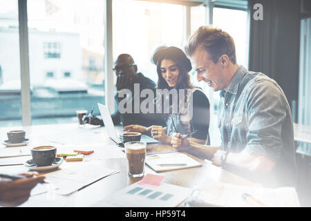 Diverse multiracial young business team in a high key office sitting working on a laptop and paperwork with happy smiles Stock Photo