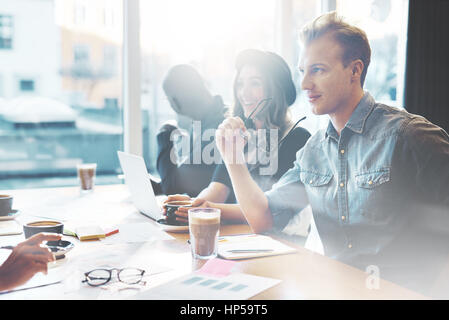 Young people talking in cafe or office, handsome man and pretty girl in casual wear having conversation with colleagues, sitting with papers and lapto Stock Photo