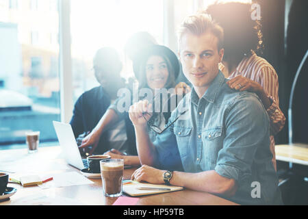 Selfconfident handsome man in casual wear holding glasses looking at camera sitting in cafe or office with colleagues or friends against bright window Stock Photo