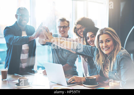 Group of young entrepreneurs in cafe giving each other high five for celebrating success or starting new project, posing and looking at camera Stock Photo