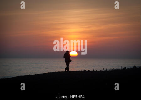 A woman stands on the beach and takes a photograph of the beautiful setting sun over the sea. Worthing, West Sussex, England, UK Stock Photo