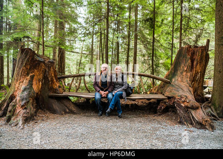 Couple sitting on hand made bench seat made from two old trees at Capilano Suspension Bridge Park Stock Photo