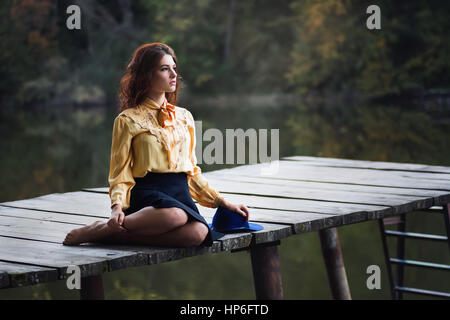 Dreaming girl with curly hair sitting on wooden bridge forest and river on background. Beautiful woman sitting by the lake. Sad girl sitting on bridge Stock Photo