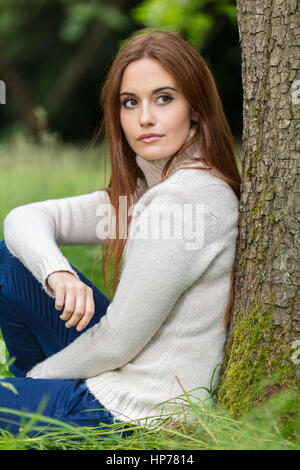 Outdoor portrait of beautiful thoughtful sad girl or young woman with red hair wearing a white jumper sitting & leaning against a tree in the countrys Stock Photo
