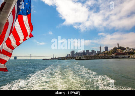 San Francisco Financial District cityscape and Oakland Bridge on sunny day, California, United States. Sea views from Alcatraz boat with American flag Stock Photo