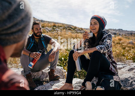 Young woman sitting with friends in countryside. Happy young hikers relaxing and having coffee. Stock Photo
