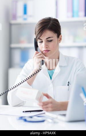 Young female doctor working at office desk, answering phone calls and reading informations on a medicine packet Stock Photo