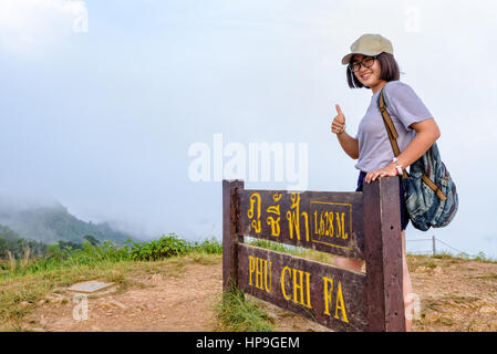 Tourist teen girl hiker wear cap and glasses with backpack smiling thumb up near nameplate attractions of Phu Chi Fa Forest Park on mountain and mist  Stock Photo