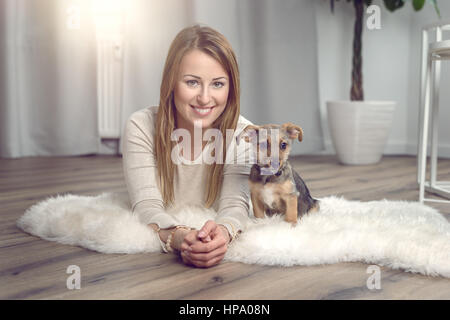 Attractive friendly woman posing with her small dog on a fluffy rug on a wood floor in the living room smiling happily at the camera Stock Photo