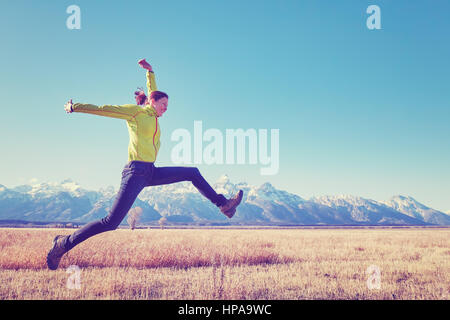 Color toned image of happy young woman jumping on a meadow, Grand Teton mountain range in distance, Wyoming, USA. Stock Photo