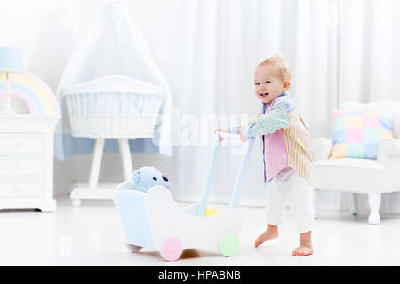 Baby boy learning to walk with wooden push walker in white bedroom with pastel rainbow color toys. Aid toy for child first steps. Toddler kid walking  Stock Photo