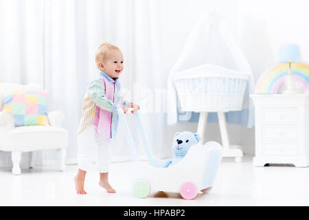 Baby boy learning to walk with wooden push walker in white bedroom with pastel rainbow color toys. Aid toy for child first steps. Toddler kid walking  Stock Photo