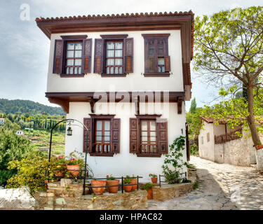 A charming traditional Turkish house with wooden shutters and intricate details sits on a stone-paved street, surrounded by potted plants and greener Stock Photo