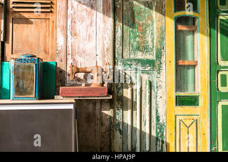 Multi-colored wall decorated with shabby wooden doors. There is a stand with an old box with mirror and a rusted sewing machine. Sunlight shines from  Stock Photo
