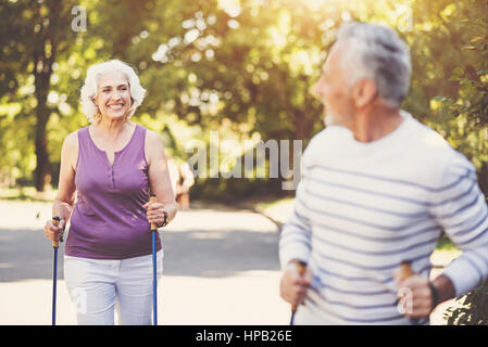 Sports activities. Joyful delighted aged woman holding the walking poles and looking at her husband while going after him Stock Photo