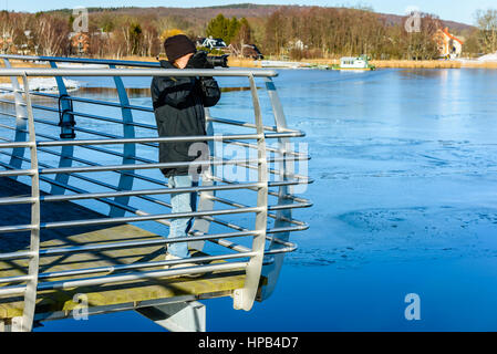 Young photographer taking picture while standing on bridge with icy water down below. Cold but sunny day in early spring or winter. Landscape in backg Stock Photo