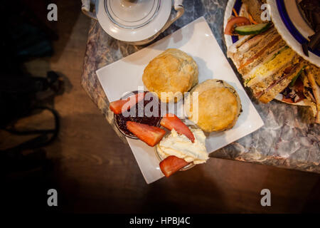 Afternoon High Tea. An overhead image of two scones on a square white plate with cream, jam and cut strawberries.  A teapot and sandwiches are nearby. Stock Photo