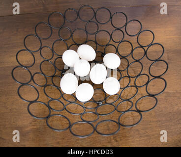 All eight white hen eggs in one black metal basket made from metal circles on a medium brown wooden table. Photographed from above. Stock Photo