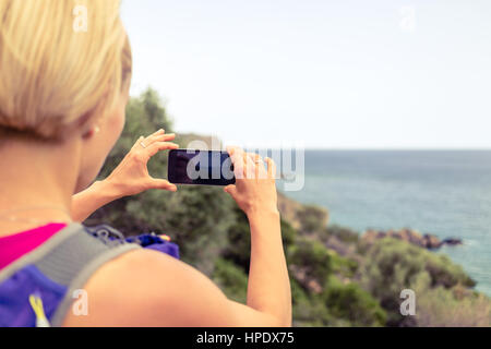 Running woman with backpack looking at inspirational summer coast, taking photo with smartphone. Travel and healthy lifestyle outdoors in nature. Fema Stock Photo