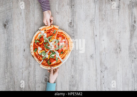 Friends sharing a delicious pizza on a rustic table, they are holding a slice, top view Stock Photo