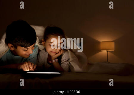 Brother and sister sharing digital tablet under blanket at night in bedroom