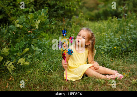 Cute little girl holding a pinwheel. She sits on the lawn. A walk in the summer park. Summer vacation. Outdoor Activities. Stock Photo