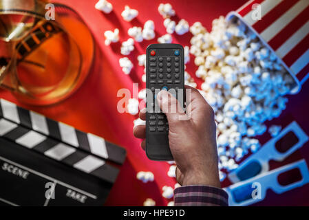 Hand holding a television remote control, popcorn, filmstrip, 3D glasses and clapper on the background, cinema and entertainment concept Stock Photo