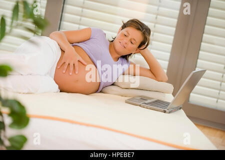 Model released , Hochschwangere Frau liegt gemuetlich am Bett, Laptop steht daneben - pregnant woman with laptop relaxing in bed Stock Photo