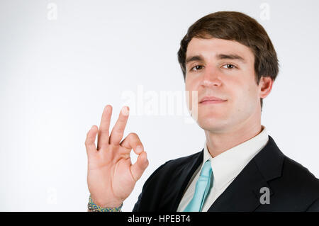 Head-and-shoulder view of a young man to dark suit against white background showing the OK sign and friendly smiling into the camera. Stock Photo