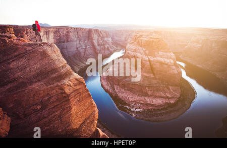 A male hiker is standing on steep cliffs enjoying the beautiful view of Colorado river flowing at famous Horseshoe Bend overlook at sunset, Arizona Stock Photo
